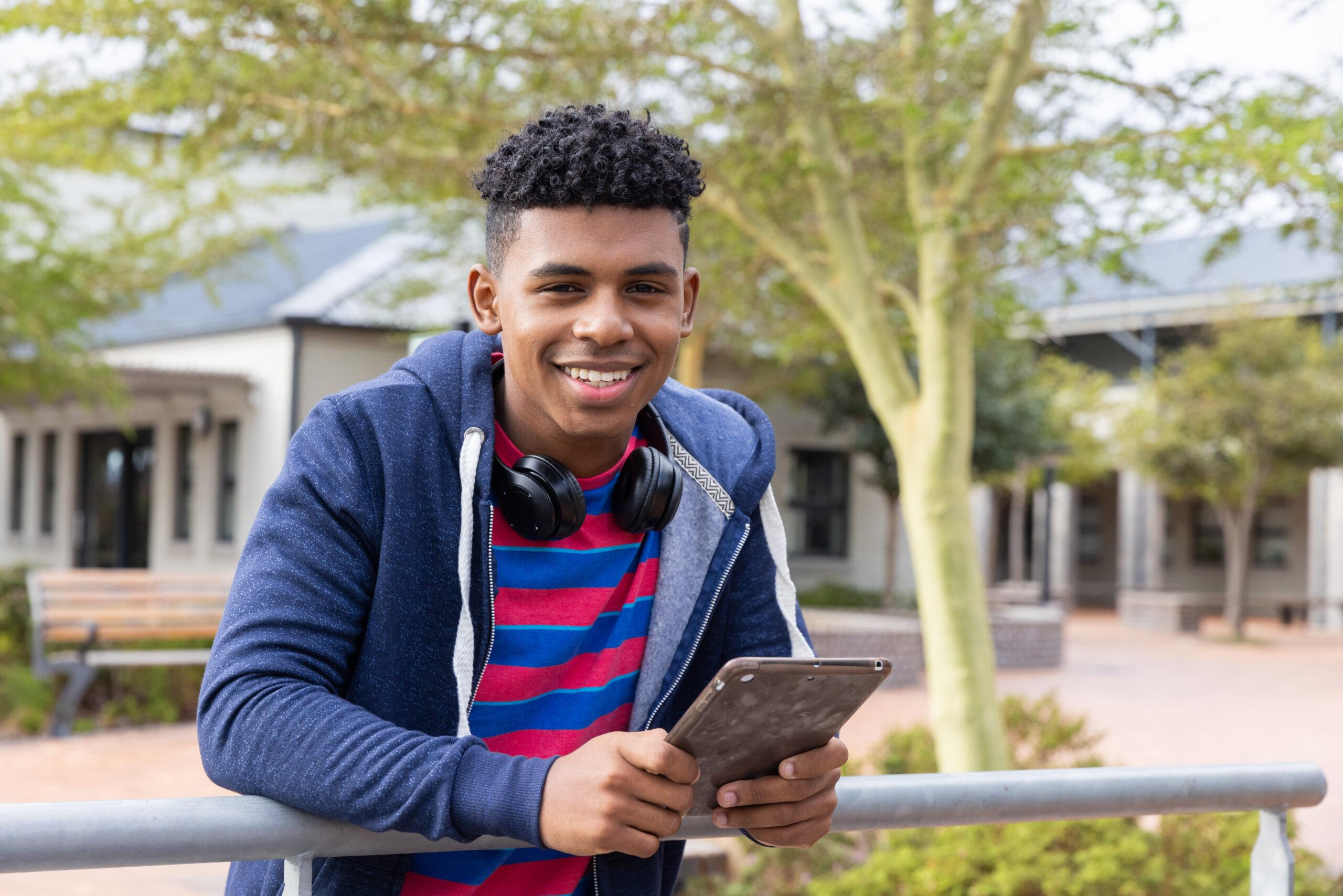 Teenage boy holding tablet, smiling and relaxing outdoors at school campus. Technology, relaxation, male student, unaltered